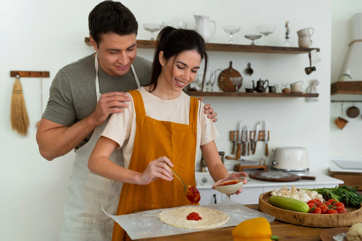 A man and a woman in a bright kitchen preparing food together. The woman wears a mustard yellow apron over a cream shirt and is spreading tomato sauce on a flatbread, while the man stands behind her with his hands on her shoulders.